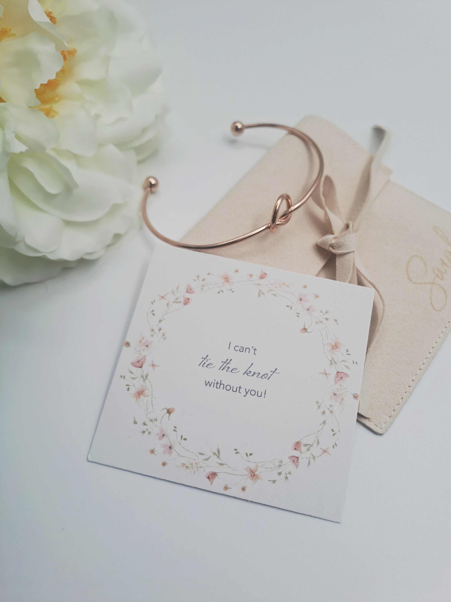 Gold bracelet on a card with floral design and text, next to white flowers on a light background