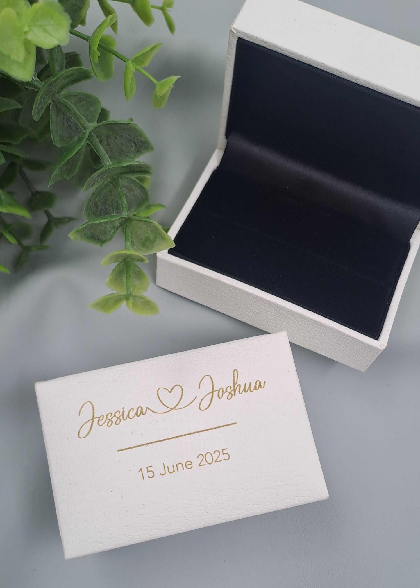 White leatherette wedding ring box with personalized gold script names and date, designed to hold two rings securely.