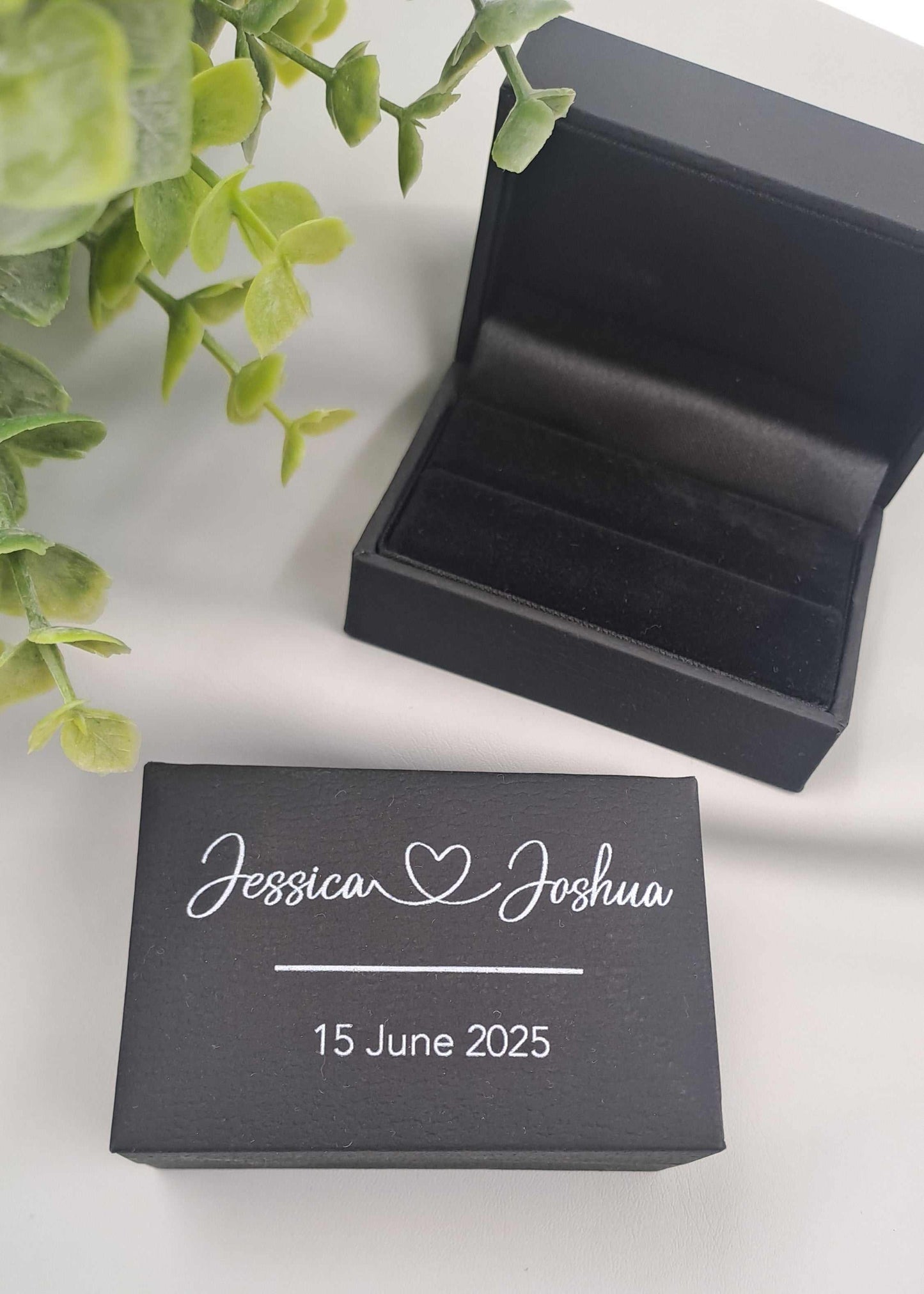 Black jewelry box with personalized name and date on a white surface with green leaves.