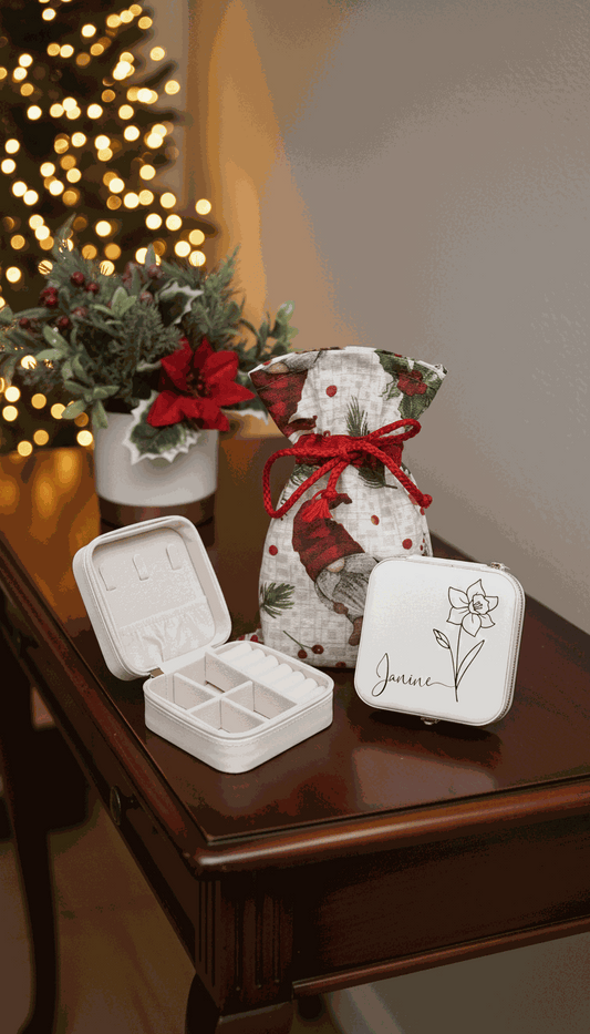 Decorative gift bag with red ribbon, jewelry box, and small white jewellery box on a wooden surface with a blurred Christmas tree in the background.