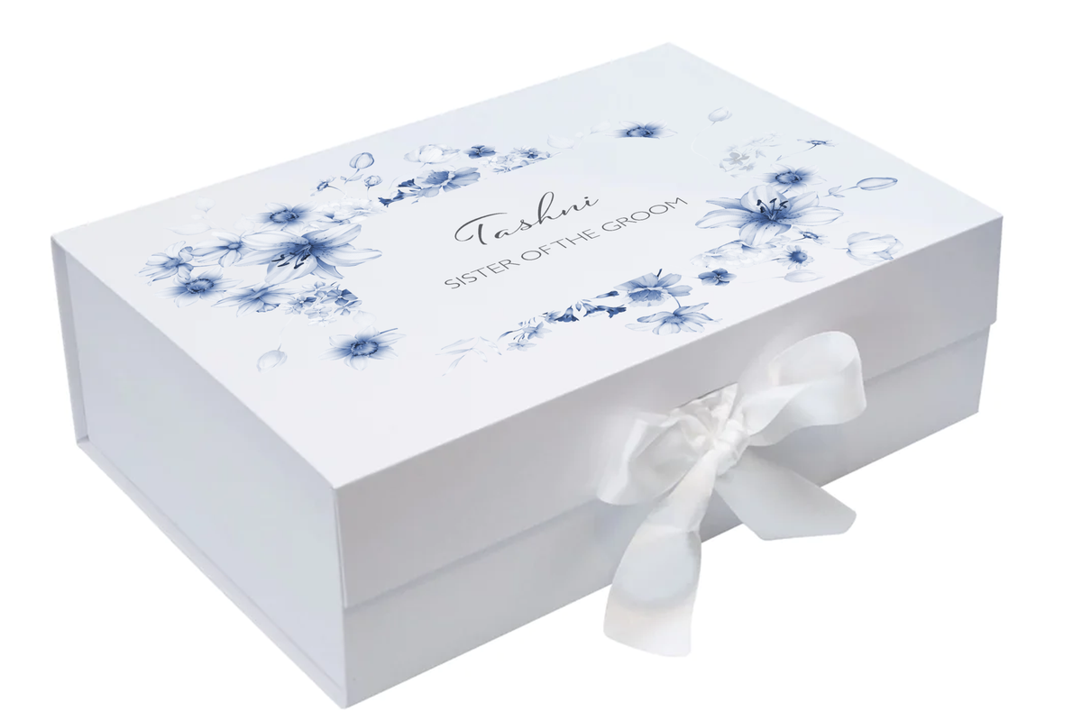 A white rectangular gift box with blue floral illustrations and the text 'Tashani SISTER OF THE GROOM' on the lid, tied with a white satin ribbon bow on a white background.