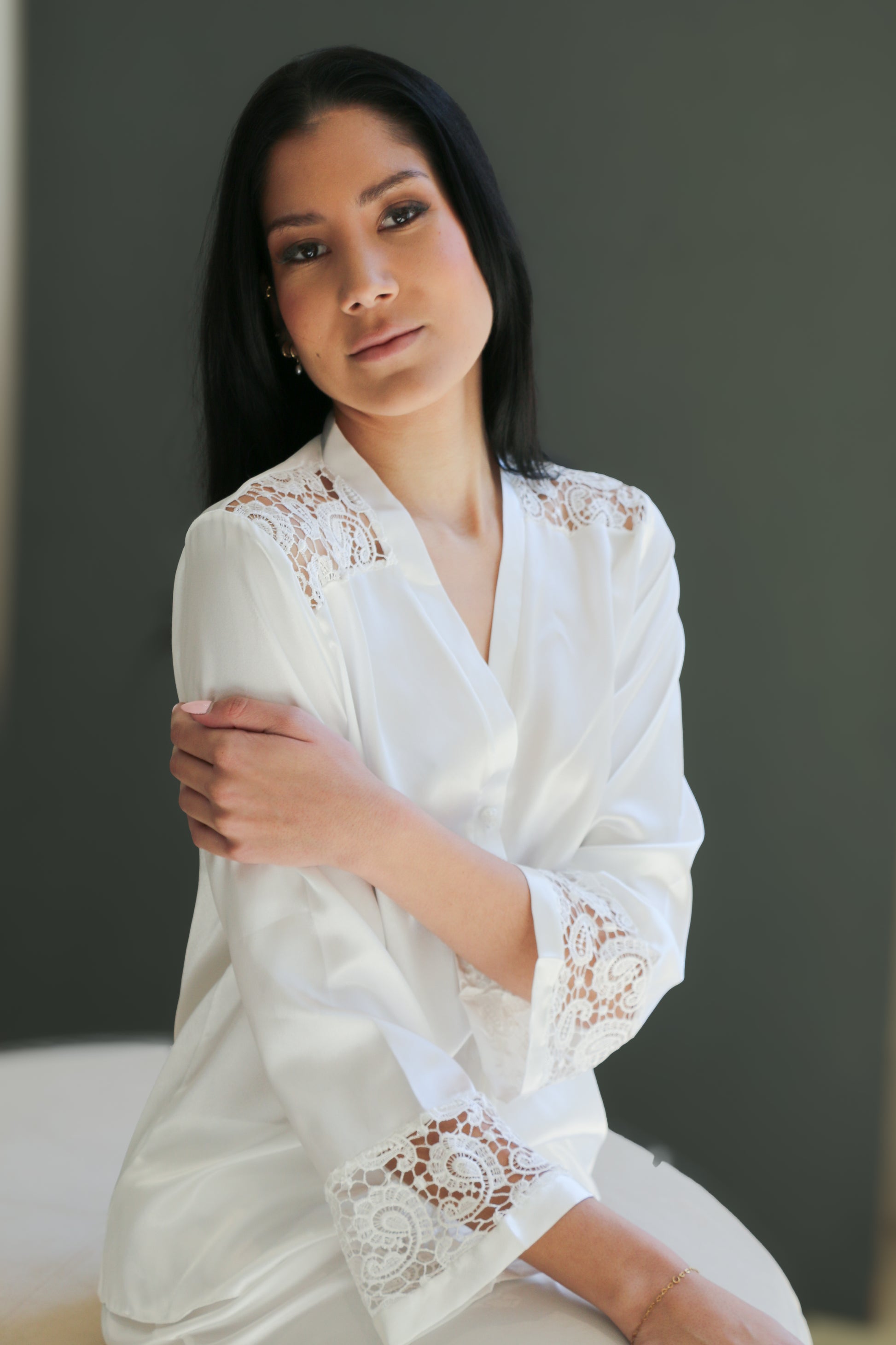 A woman wearing a white satin-like robe with intricate lace embroidery on the shoulders and cuffs, posed against a dark gray background.