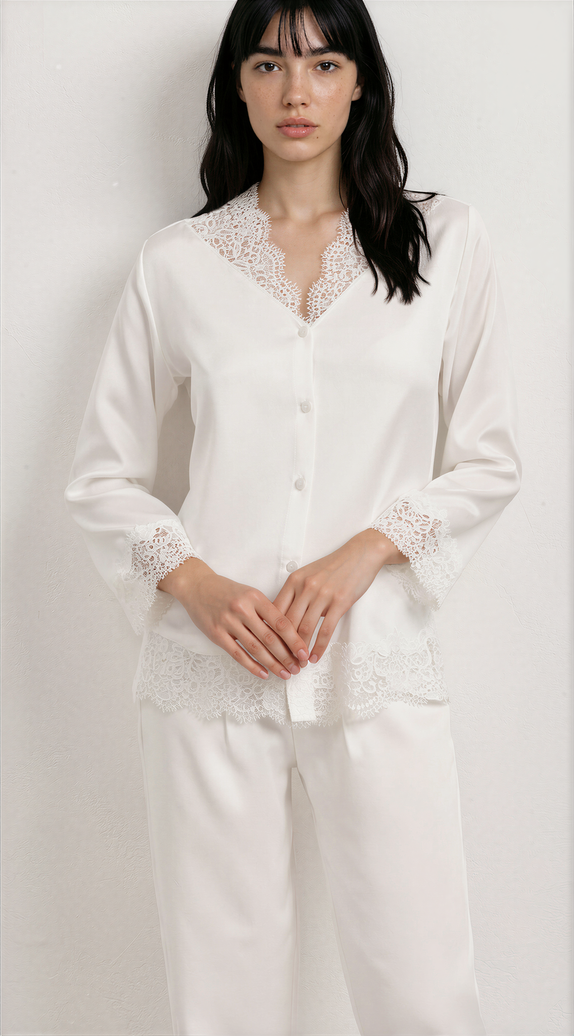 A woman modeling a white long-sleeve pajama set with delicate lace trim along the V-neckline, cuffs, and hem, paired with matching white pants against a plain light background.