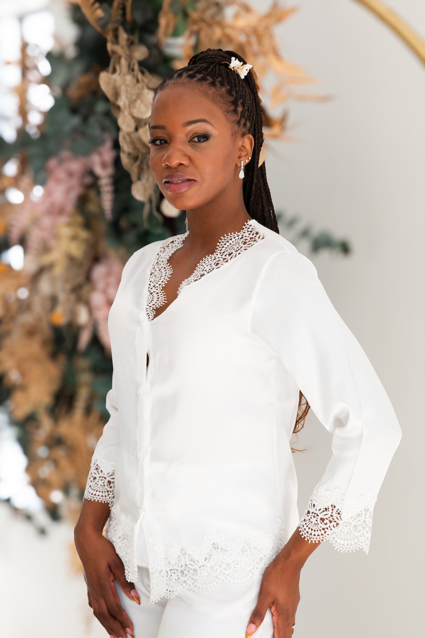 A woman wearing a white long-sleeve blouse with intricate lace trim at the V-neck, cuffs, and hem, paired with matching white pants. She is posed in front of a soft-focus floral and boho decor background.
