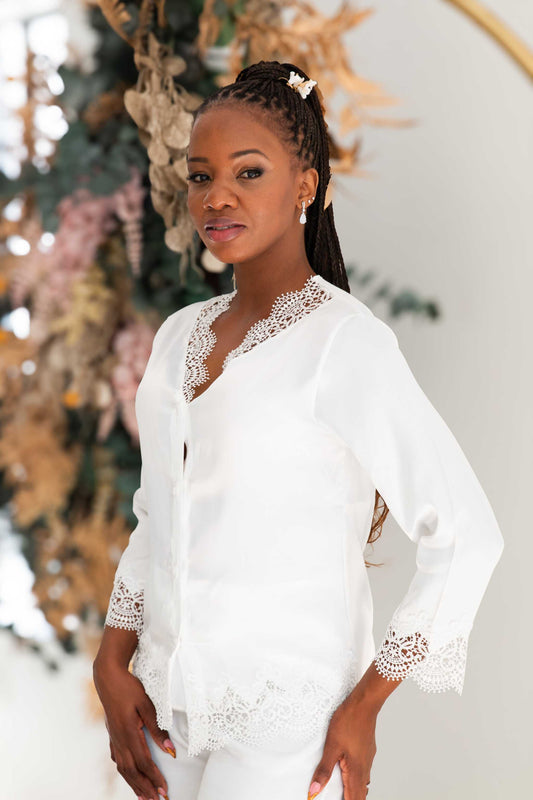 A woman wearing a white long-sleeve blouse with intricate lace trim at the V-neck, cuffs, and hem, paired with matching white pants. She is posed in front of a soft-focus floral and boho decor background.